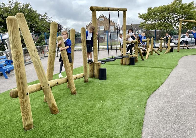 A class of children playing on their trim trail, artificial grass surrounds this area. The children are playing with the trim trail in a single file line.