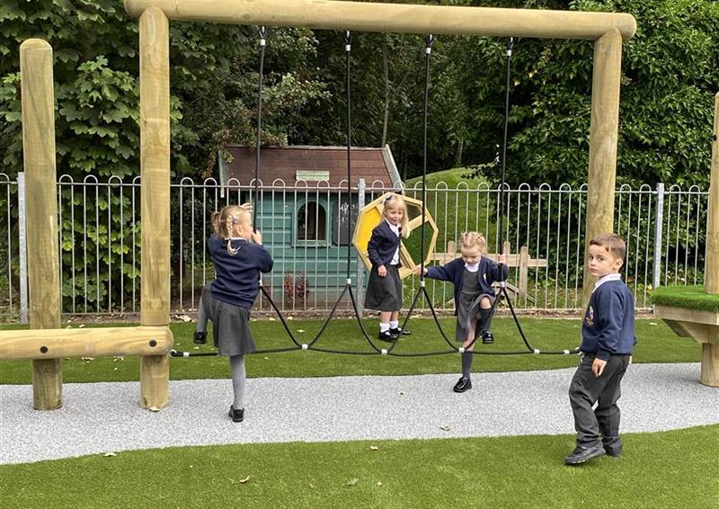 3 children playing on their trim trail, 1 young boy is playing on the side. A layer of artificial grass can be seen on both sides of the Trim Trail, as a thin strip of grey wetpour is laid underneath the trim trail.