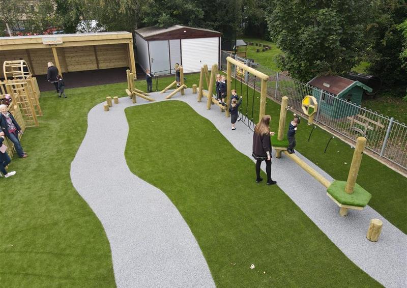 A birds eye view of a class of children playing on their trim trail, 1 teacher is stood to the side accompanying the children. Artificial grass covers the play area, with a wetpour road design placed underneath the Trim Trail itself.