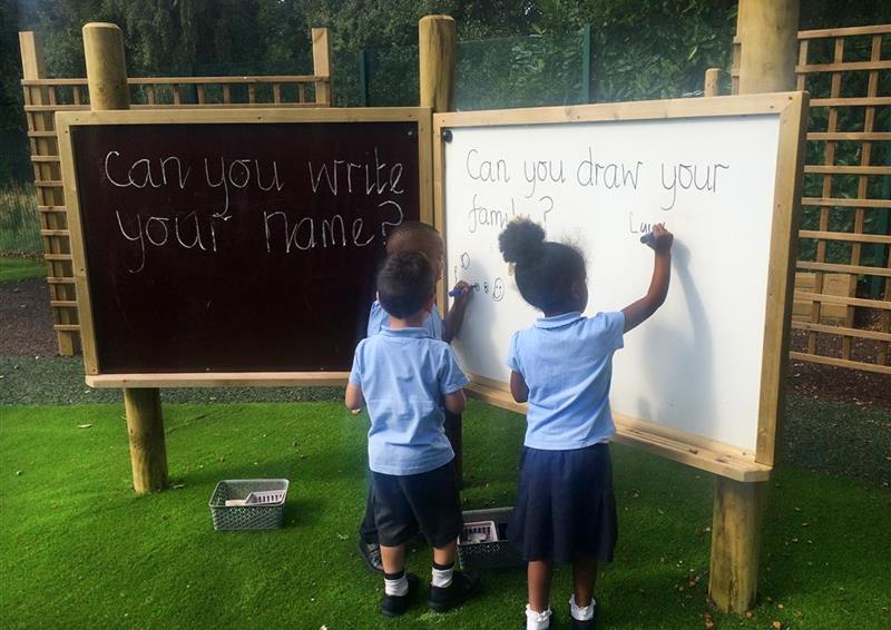 3 children drawing on a giant white board with the giant chalk board next to them. 2 of the children are writing and one of the children on watching.