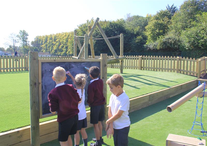 3 children drawing on a giant chalk board, one young boy is walking behind them.