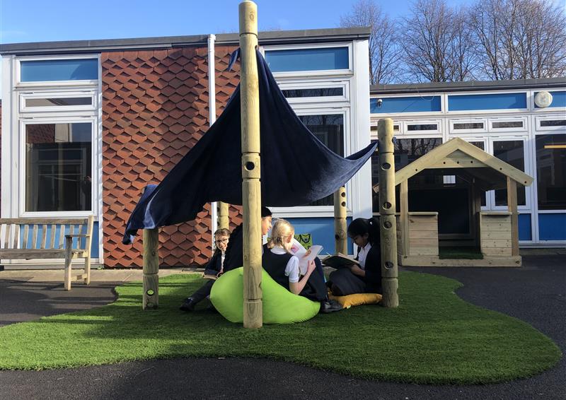 4 children sat underneath the den making post, they are sat on bean bags with a sheet on top of the den making posts creating a shelter.