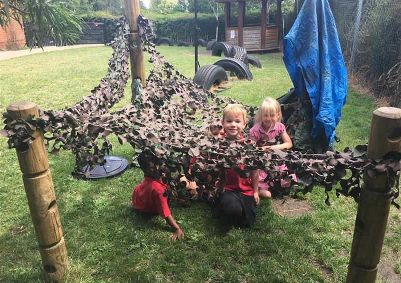 3 children playing underneath their den making posts, they have used a leaf net over the top to create a shelter.