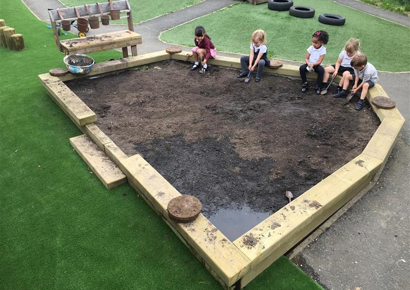 children playing in their dug out pit which is full of mud, the children are sat around the edge of the sleeper and digging. The surface the Dig Pit is installed on is artificial grass.