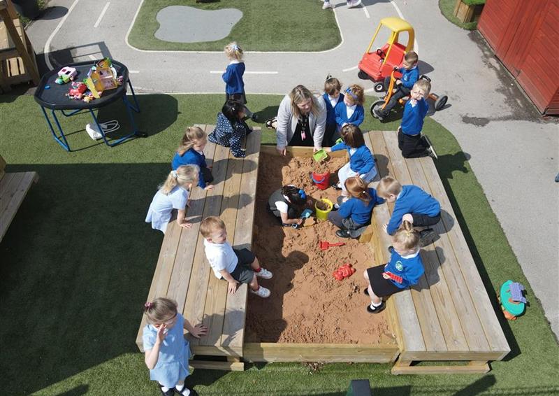 A class of children and a teacher are all playing in the sand box, they are digging and making sand castles.