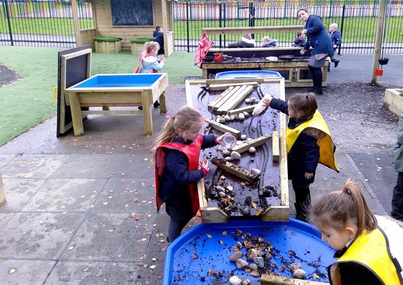 3 children playing on their damming station, they are using bottles of water to do their experiment, there is rocks and pieces of wood to act as the dam