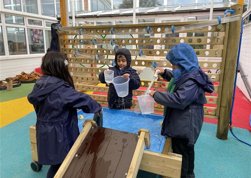 3 children playing on the fine motor table, they are all wearing rain coats and using jugs to fill water and pour down the damming station.
