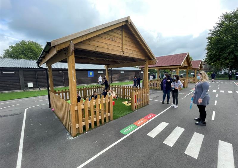 timber shelter with bow top timber fencing and artificial grass surfacing below with playground markings showing a roadway whilst three staff members looking into it