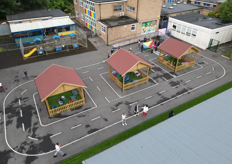a birdseye view of three bespoke timber canopies on a playground with artificial grass surfacing within and a playground marking of a roadway around the perimeter