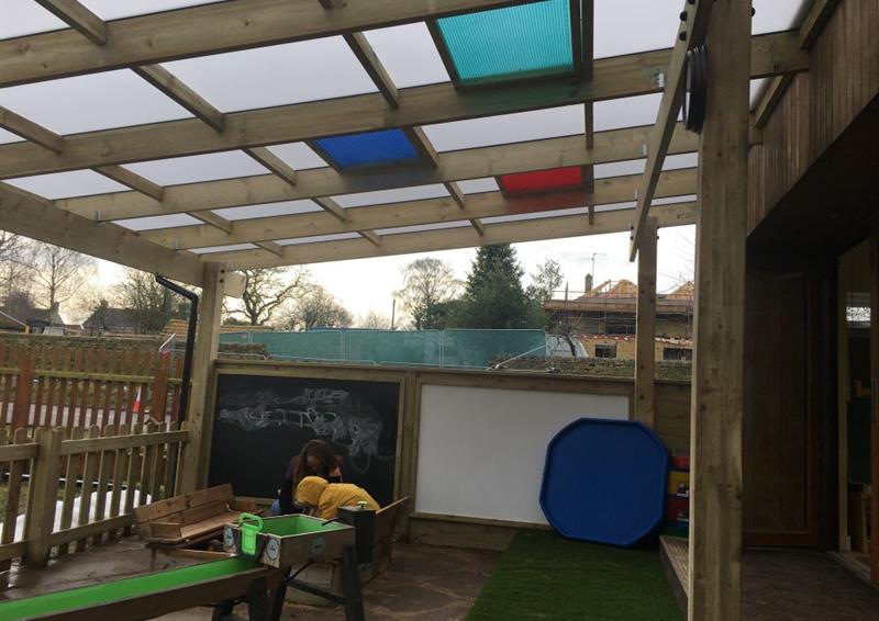 a timber canopy with red, green and blue panels fitted into the roof and a play area with artificial grass and a tuff spot table underneath