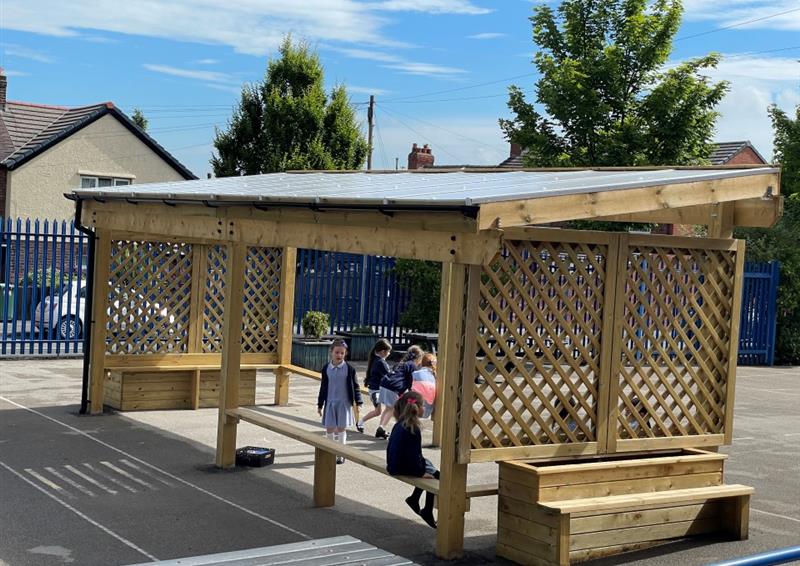 Canopy with a timber trellis sides and bench inside with people sat on it reading, there is planter benches on the outside