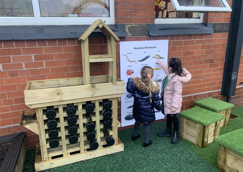 2 girls looking at their identifying bug board next to their bug hotel, there are grass top seats scattered around.