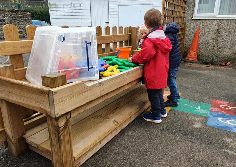 2 young boys playing on their construction table which has toys in. The table has been placed on a tarmac playground, with a thermoplastic playground marking next to the table. A little girl is at the end of the table, playing with the equipment.