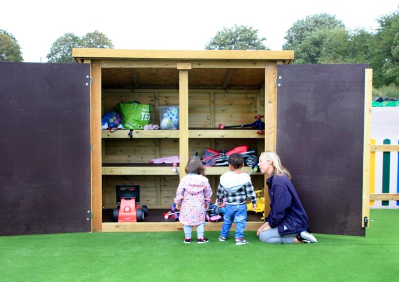 an image showing two nursery age children and a teacher standing in front of the large store cupboard that is filled with classroom resources, there is artificial grass beneath them