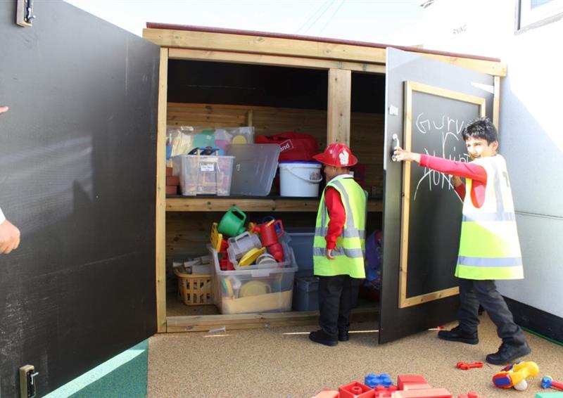 two children in red school uniforms and hi vis vests looking into the timber store cupboard with chalkboards on the doors