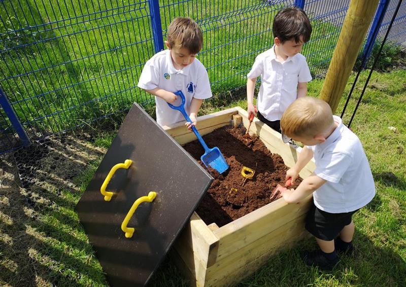 3 children playing in their mud box, there are spades and toys within the box. The Mud Box has been placed on a natural grass turf, with the boys looking in the box.