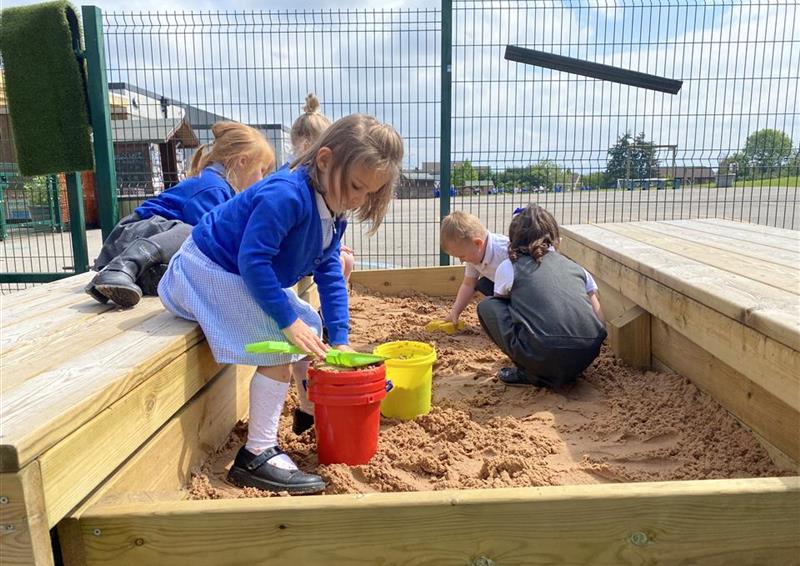 3 children playing in their large covered sandbox, they are building sand castles. The sand box has been installed on top of concrete with a green fence in the background.