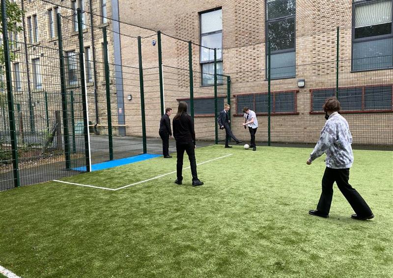 4 children playing on the MUGA which has a blue goal and netting on top. The surface is artificial grass and the MUGA is placed right next to the school building.