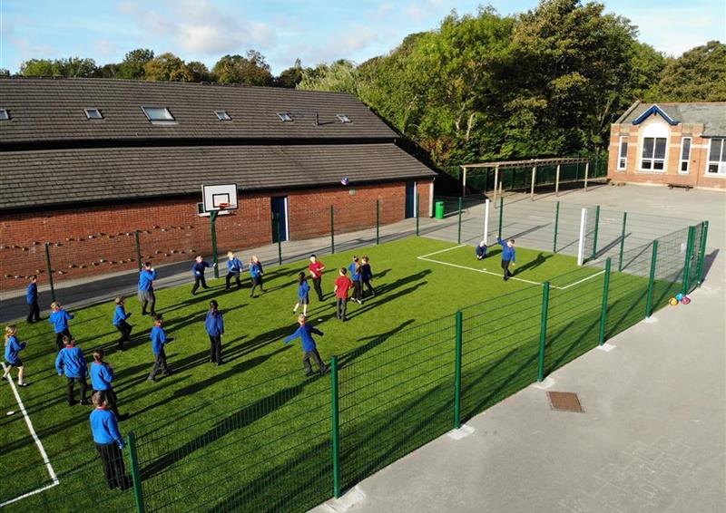 A class of children playing football on their multi-use games area. The MUJGA has a green fence going around the surface, with a football goal being seen on both sides. A basketball hoop is attached to the fence, allowing for basketball.