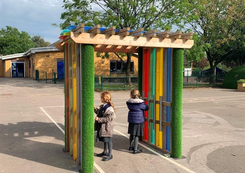 a colourful sensory arbour with red, yellow, blue and green colourful panels with two children playing on the inside wearing winter coats