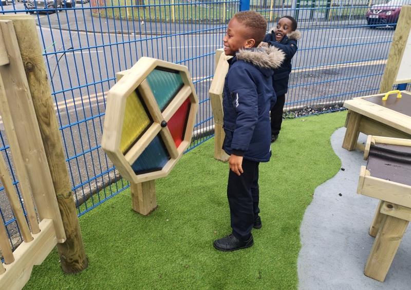 a child in a navy blue winter coat with a fur hood stands and looks at the sensory spinner which is made from timber and multi-coloured polycarbonate panels