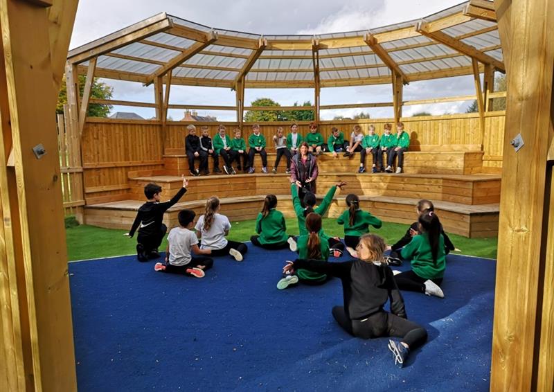 inside one of our musical ampitheatres with a performance stage and seating, children sit on the benches and watch their peers as they sit on the performance stage talking to them