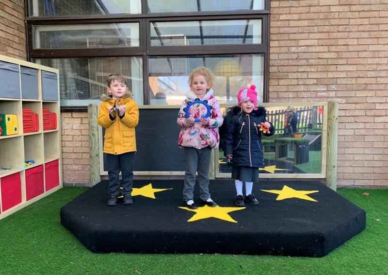 three children in winter coats and hats pose on the black performance stage with felt stars on the surfacing and a giant mirror and chalkboard in the back