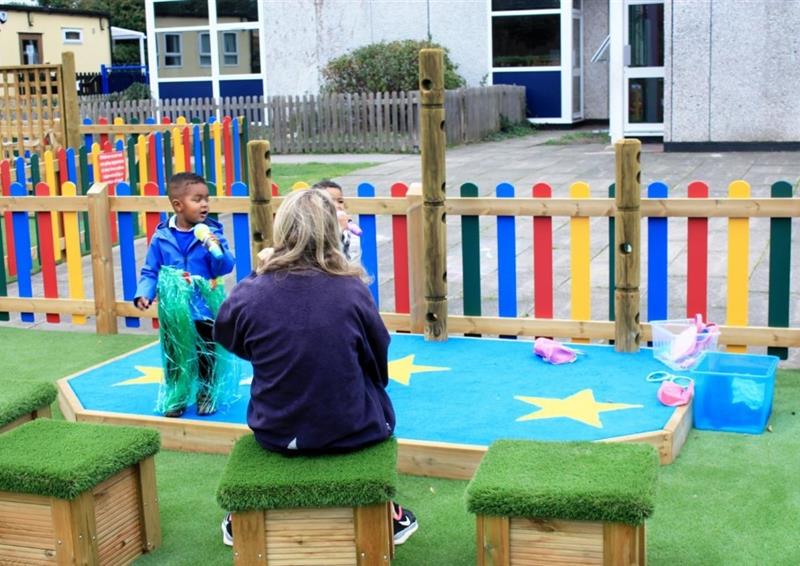a teacher sits on artificial grass-topped seats and watches a child in a winter coat perform on the blue performance stage with rainbow fencing behind