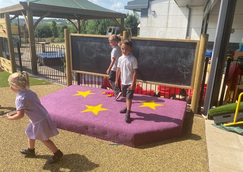 a purple surfaced performance stage with a giant chalkboard behind with three children dancing and performing on the stage