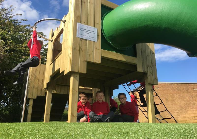 3 children playing around the langley play castle, 2 children are sat underneath and 1 child is swinging from the fireman's pole