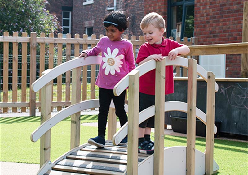 Two EYFS school children walking over play bridge. The play bridge is wooden and has been placed on an artificial grass surface. The school building can be seen in the background.