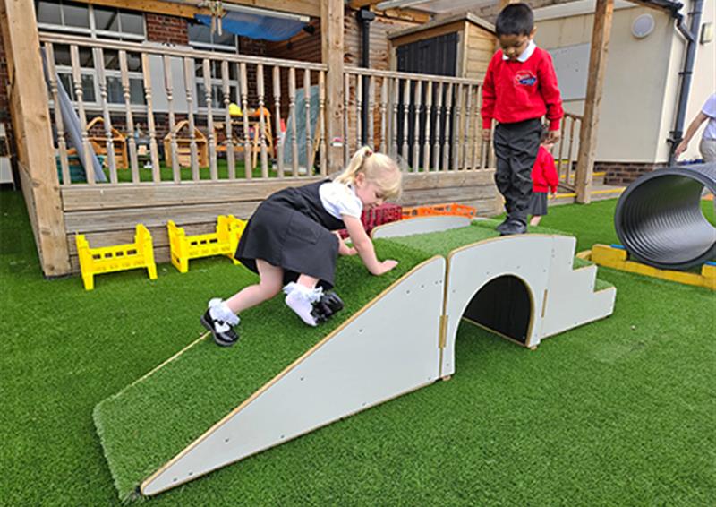 School children playing on up and under blocks, which have been placed on an artificial grass surface. One child is crawling up the ramp, whilst the other child is climbing up the stairs, meeting in the middle.