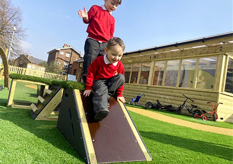 School children playing on Get Set Go Blocks, which have been placed on artificial grass. One child is sliding down a slide block whilst the other child is stood on the top of the block, looking down the slide.