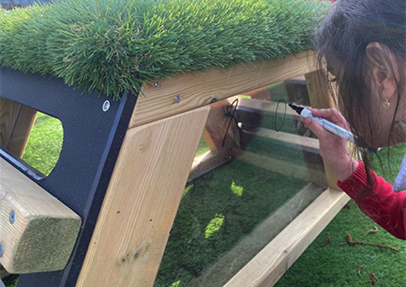 A school child drawing on clear panel on the Get Set, Go Block with a black marker. The top of the Get Set, Go! Block is artificial grass, with the clear panel being surrounded by natural wood.