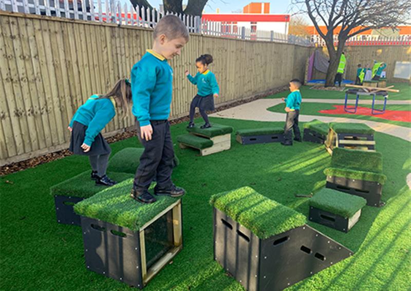 School children moving from one Get Set, Go! Block to others. The obstacle course has been placed on an artificial grass surface, with a path behind the course leading to another play area.