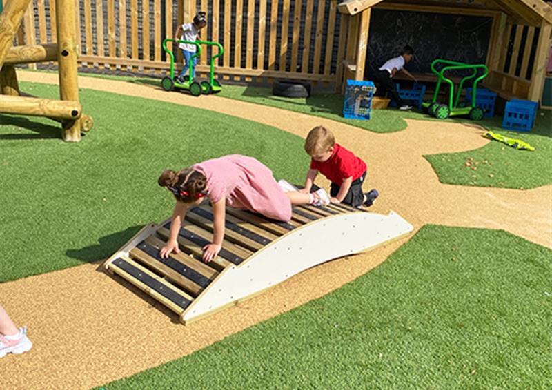 School and nursery children climbing on the Play Bridge. The play bridge has been placed on a wetpour path, with artificial grass surrounding the path. The children are crawling on behind the other over the bridge.